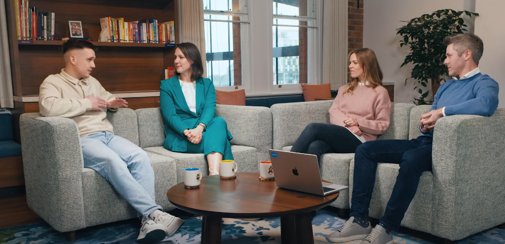 Four people are having a conversation while seated on grey couches in an office.